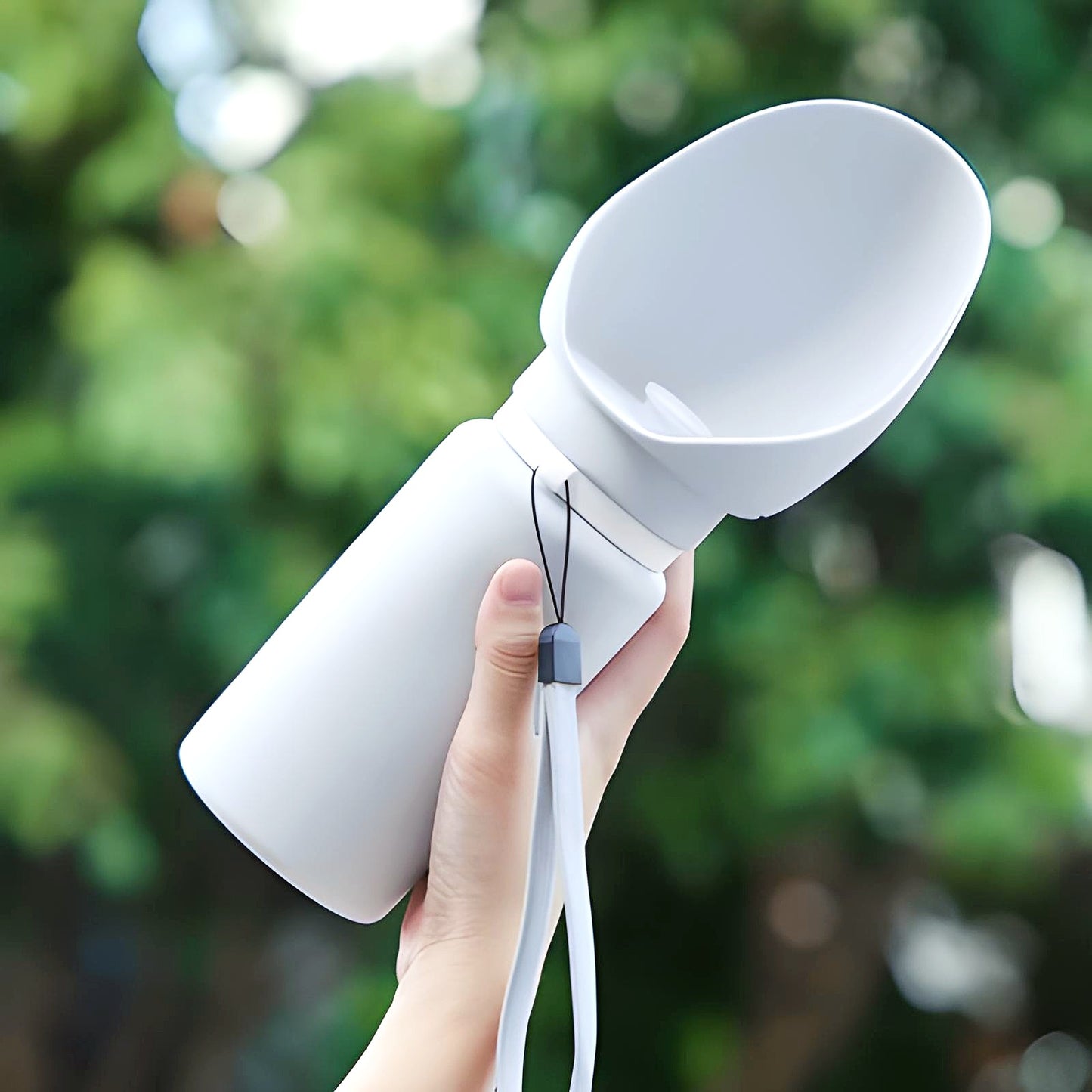 White 550 ml Sip-n-Stroll™ portable dog water bottle held upright with wrist strap against a green outdoor background